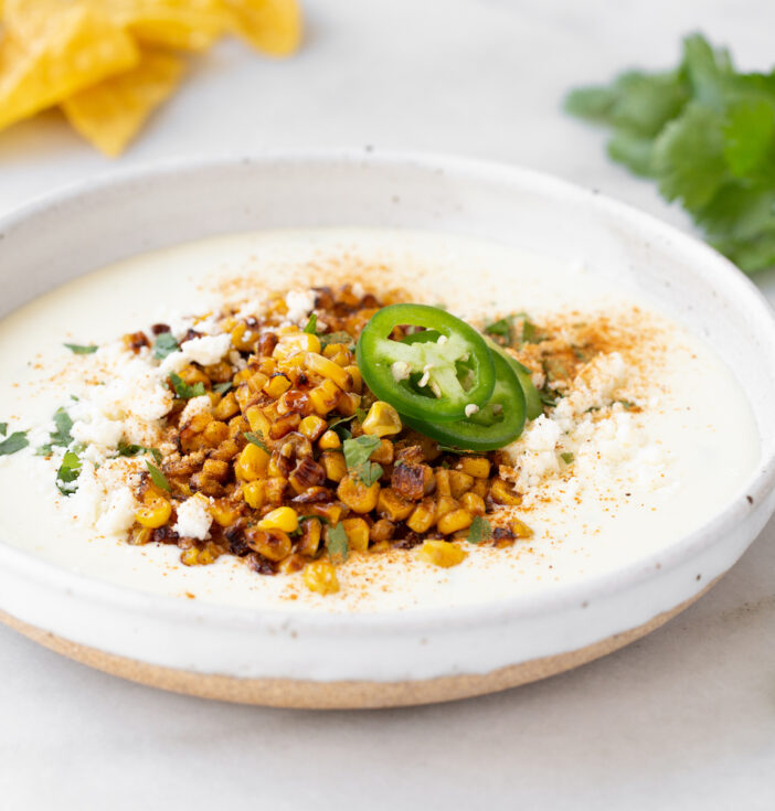 A white bowl of dip topped with corn and jalapeños