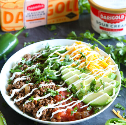 A taco salad with a white dish surrounded by peppers, sour cream and a block of cheese