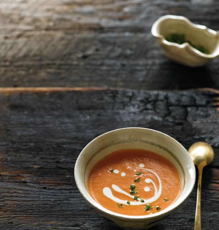 A small bowl of tomato soup displayed on a slab of wood with a brass spoon