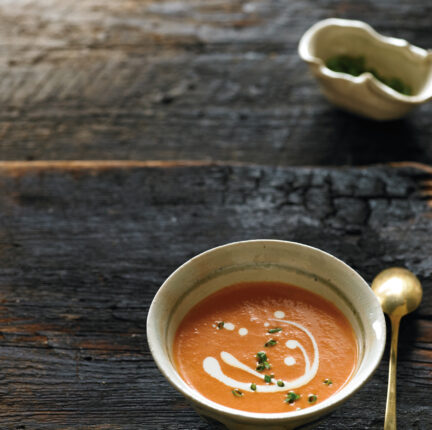 A small bowl of tomato soup displayed on a slab of wood with a brass spoon