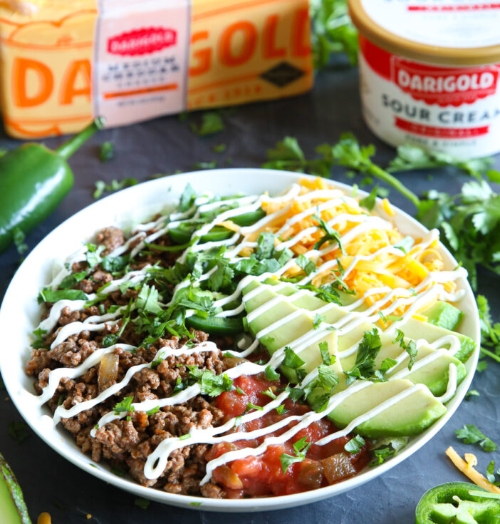 A taco salad with a white dish surrounded by peppers, sour cream and a block of cheese