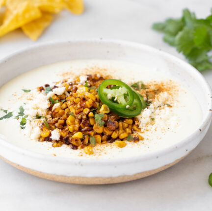A white bowl of dip topped with corn and jalapeños